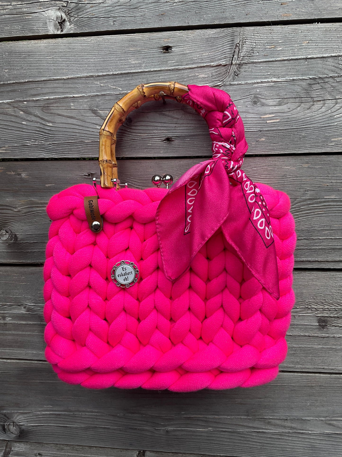 Eine gestrickte pinke Trachtentasche mi einem um den Griff geknotetem Tuch und einer Brosche.