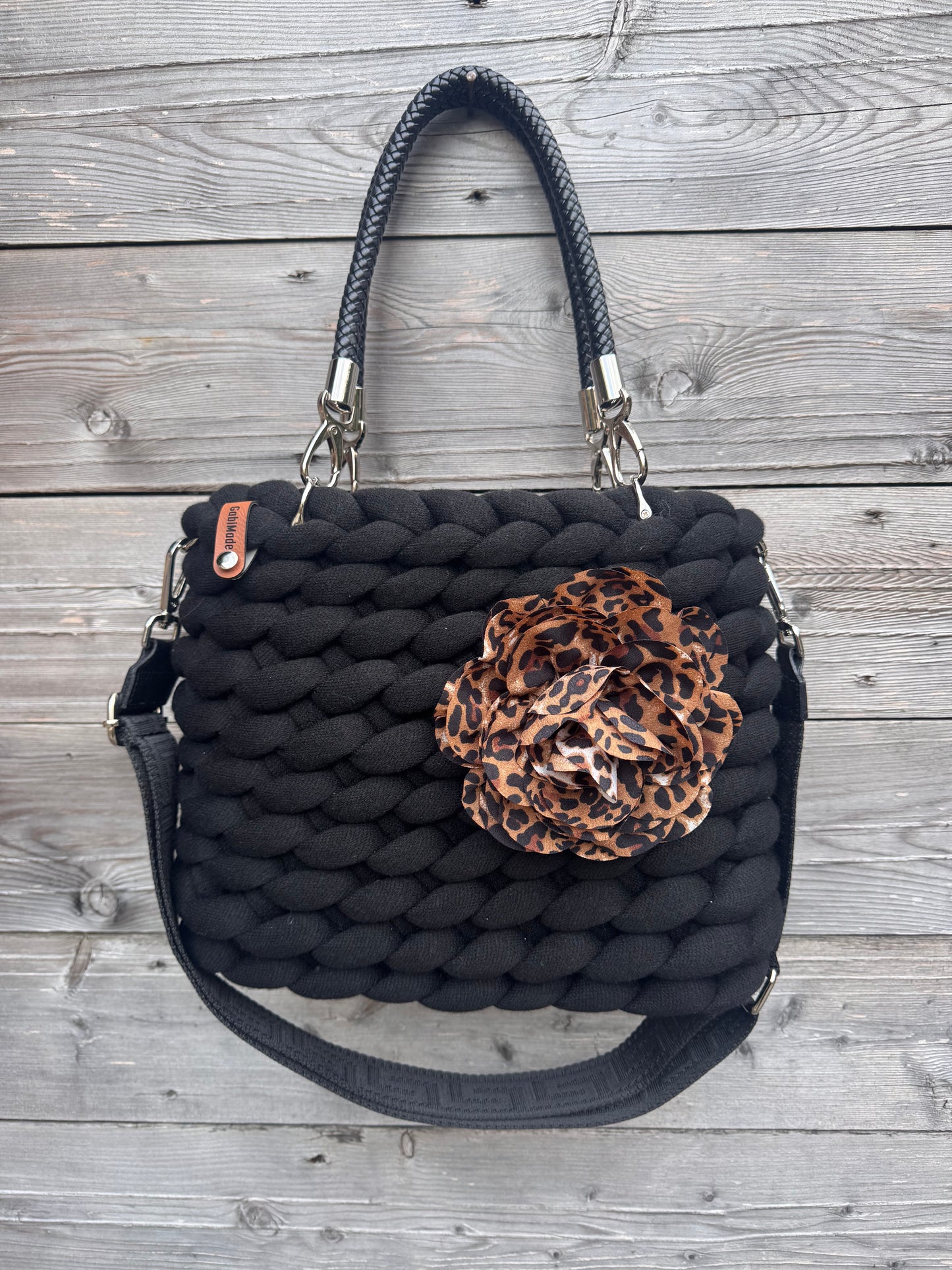 Eine gestrickte schwarze Tasche mit schwarzen Griffen, Blumenbrosche und Gurt